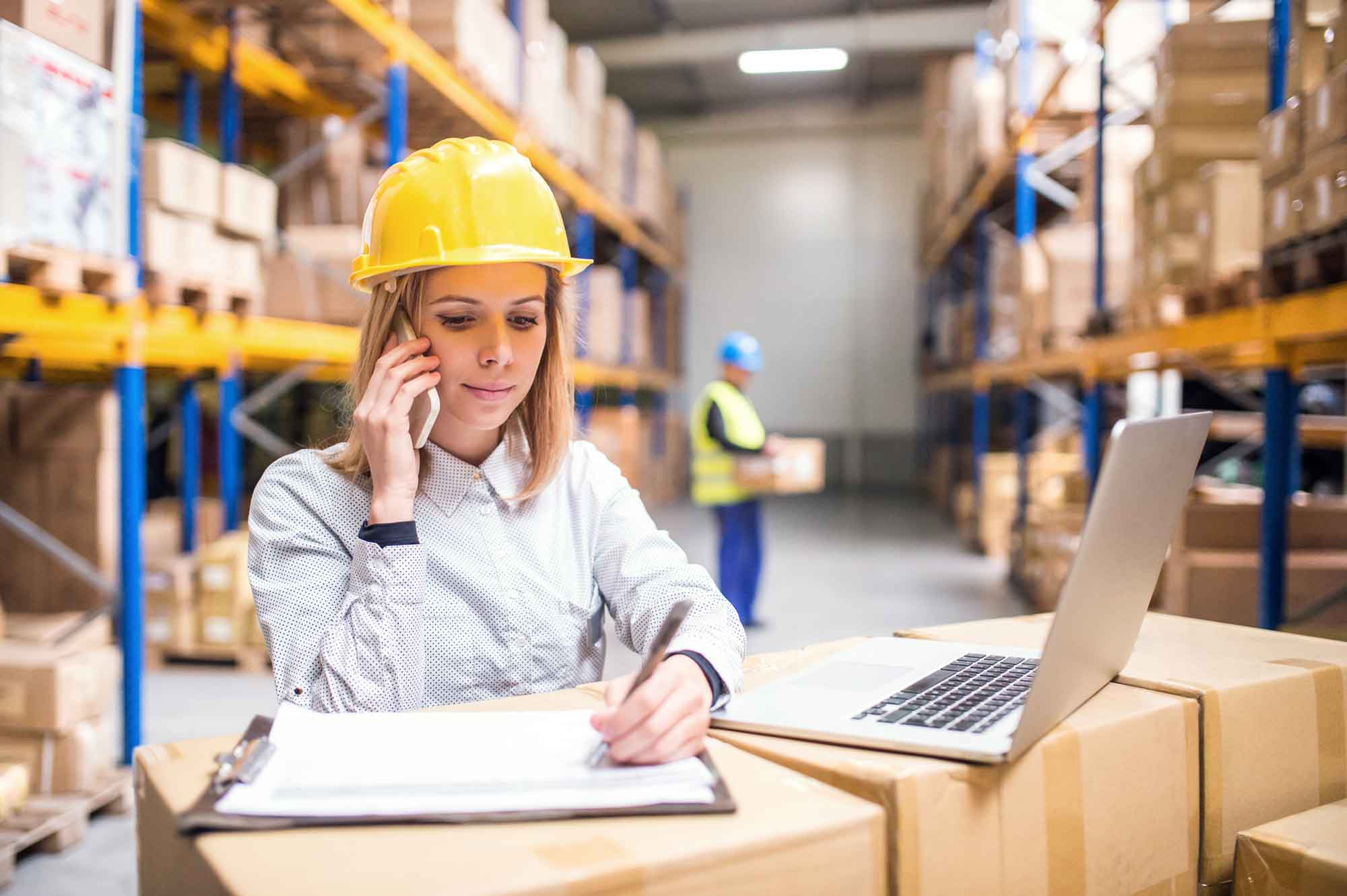 Certificat Supply Chain e-commerce, formation continue de Dauphine Executive Education (visuel objectifs) Une professionnelle de la supply chain travaille avec un smartphone et un ordinateur dans un entrepôt rempli de cartons. Programmes de formation continue en Supply Chain et e-commerce de Dauphine Executive Education
